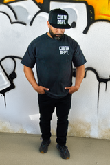 Person wearing a black 'CULTR DEPT.' t-shirt and cap against a graffiti-covered wall.